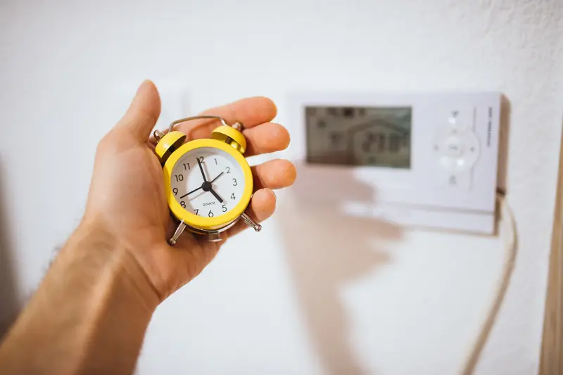 A hand holds up a yellow wind-up alarm clock beside a programmable thermostat on the wall