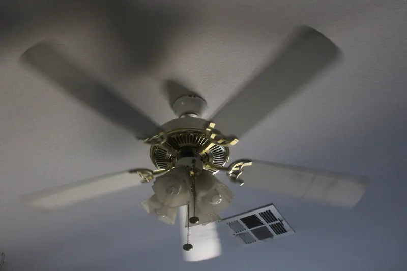 A white ceiling fan mid-spin with its blades blurred, mounted near a ceiling air vent