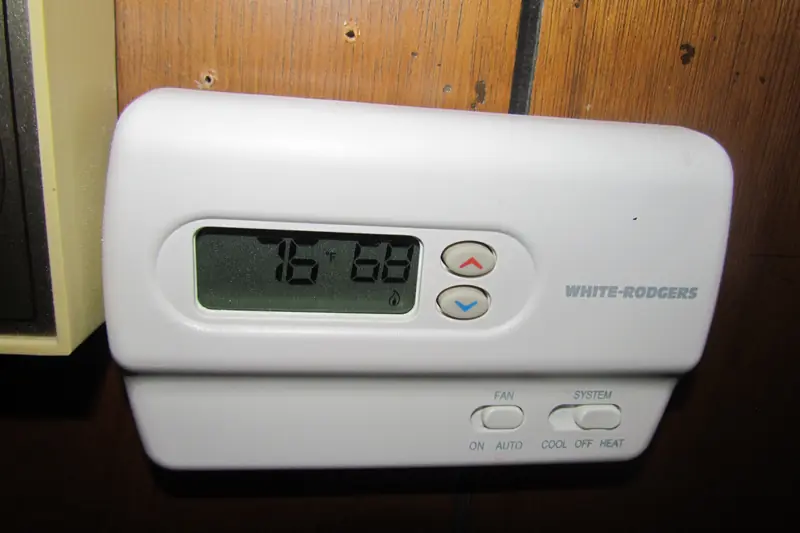 Beige wall thermostat on dark wood paneling, its screen showing 76 and 68, with sliding fan and system switches along the bottom edge