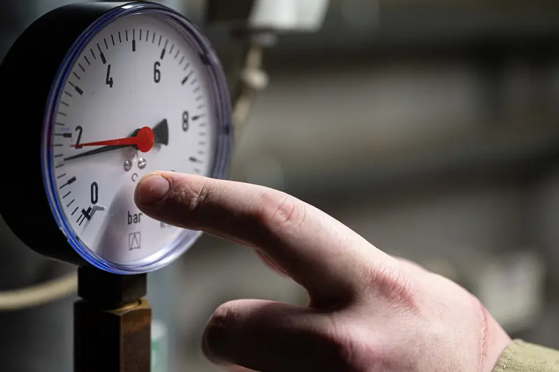 Finger pointing at the red needle of a round dial pressure gauge with black numbers, pipes blurred behind it