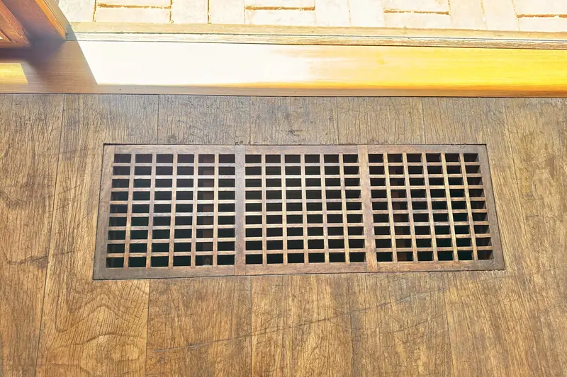 A metal floor register with rectangular louvers set into a wood floor below a window
