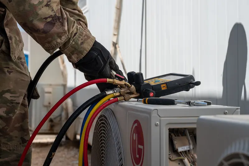 Gloved hands of a technician holding a digital manifold gauge with red, blue, yellow and black hoses clipped to a small outdoor unit