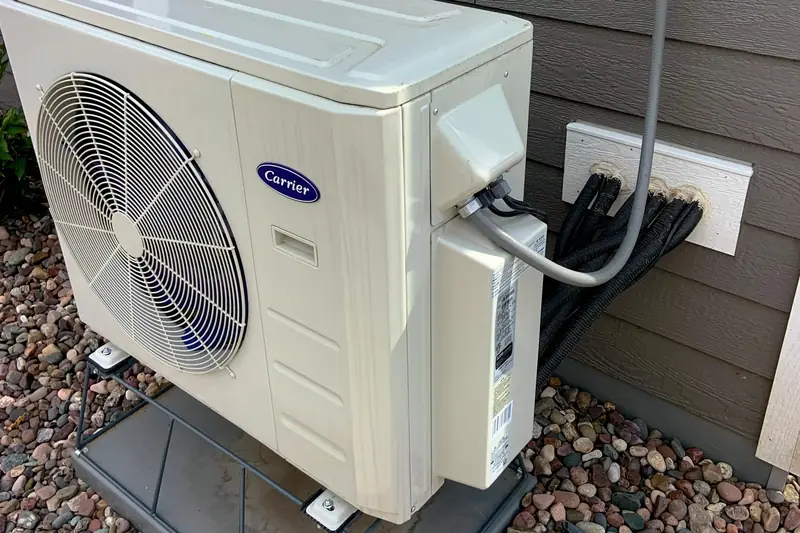 A compact mini split heat pump outdoor unit on a pad against grey siding, with its electrical disconnect box mounted above