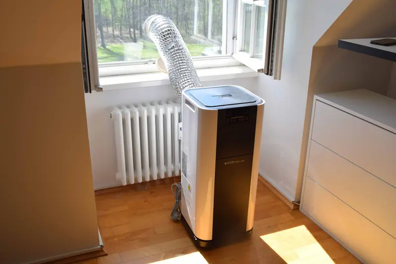 Tall white portable air conditioner on a honey-colored wood floor, a silver accordion exhaust hose rising to an open sash window beside an old radiator