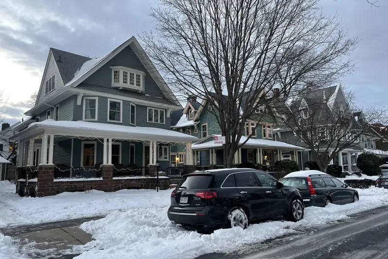 Snowy residential street of tall shingled houses with wide porches, a black SUV parked at a cleared curb and bare trees overhead